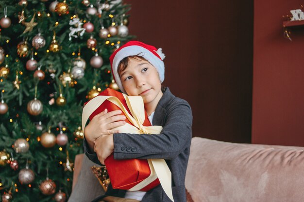 Attraktiver lustiger Tweenjunge mit dem dunklen Haar in Sankt-Hut umarmt Geschenkbox auf Weihnachtsbaum