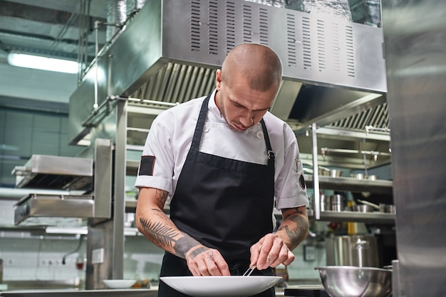 Attraktiver männlicher Koch mit Tattoos auf den Armen, der sein Gericht auf dem Teller in der Restaurantküche garniert