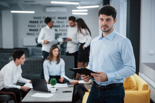 Attraktiver Manager, der bereit ist, Ihre Fragen zu beantworten. Porträt des jungen Mannes steht im Büro mit Mitarbeitern im Hintergrund