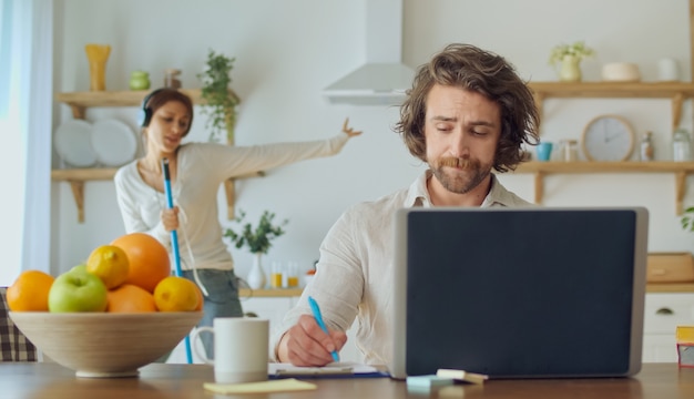 Attraktiver Mann, der versucht, aus der Ferne zu arbeiten, während seine Frau mit Kopfhörern tanzt und Karaoke singt. Frau mischt sich in die Arbeit ihres Mannes ein.