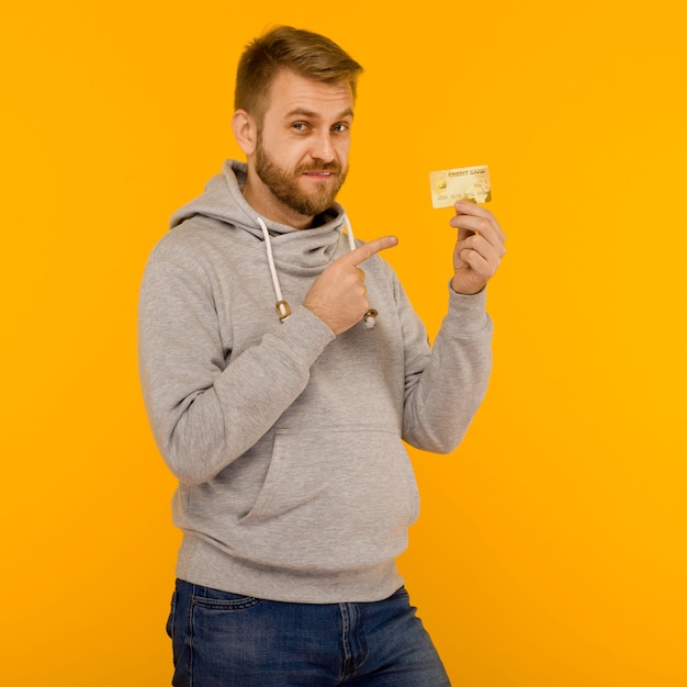 Attraktiver Mann in einem grauen Kapuzenpulli zeigt mit einem Finger auf die Kreditkarte, die in seiner Hand auf einem gelben Hintergrundbild hält