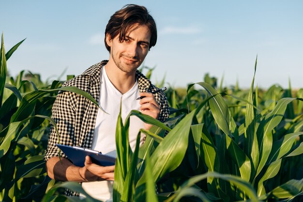 Attraktiver, selbstbewusster Mann, der in einem Maisfeld arbeitet. Der Agronom schaut in die Kamera und hält Notizen.