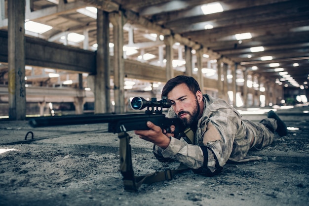 Foto attraktiver und ernster kerl liegt aus den grund in einem großen hangar. er zielt. mann beim schauen durch linse. er ist sehr ruhig und konzentriert.