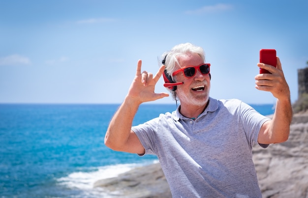 Attraktiver verrückter älterer Mann, der Selfie am Meer macht, rote Kopfhörer und Sonnenbrille trägt. Sorglos im Ruhestand Spaß mit Smartphone-Playlist-Apps