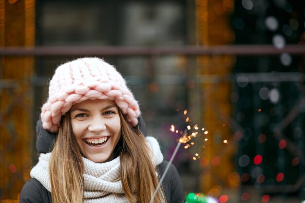 Attraktives blondes Mädchen mit Winteroutfit, das Spaß mit Wunderkerzen auf der Straße hat. Platz für Text