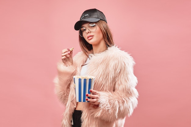 Attraktives brünettes Mädchen in rosa Kunstpelz, das Popcorn in der Hand hält und in schwarzen Shorts weißes Top trägt ...