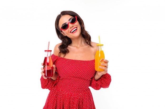 attraktives brünettes Mädchen mit Sonnenbrille im roten Kleid mit einer Cocktailbrille auf einer weißen Wand