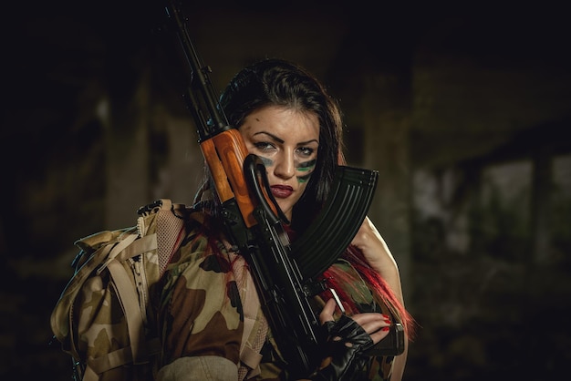 Attraktives dunkelhaariges Mädchen im Tarnhemd hält ein Maschinengewehr, das sich auf die Schulter lehnt und mit einem gefährlichen Gesichtsausdruck in die Kamera schaut.
