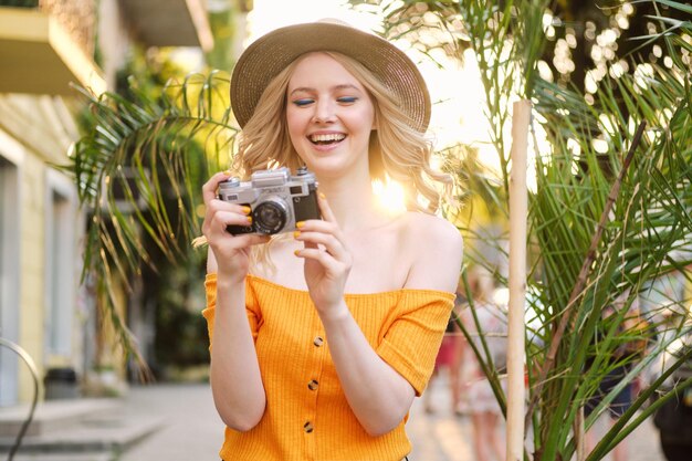 Attraktives, fröhliches blondes Mädchen mit Hut, das glücklich mit einer Retro-Kamera auf der Straße der Stadt fotografiert