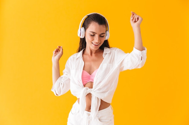 Attraktives fröhliches junges Mädchen, das Sommerstrandkleidung trägt, tanzt, während es Musik mit Kopfhörern hört, die über gelber Wand isoliert sind?