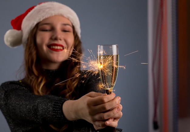 Attraktives, fröhliches Mädchen mit lockigem Haar in Weihnachtsmütze, lächelnd, Wunderkerze haltend und Champagner isoliert auf grauem Hintergrund trinkend