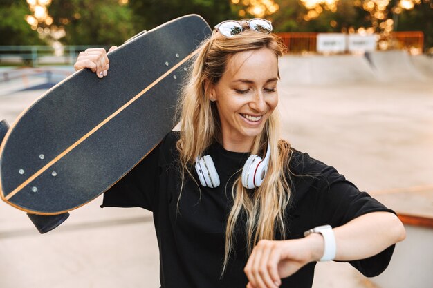 Attraktives funky Teenager-Mädchen trägt Longboard beim Gehen im Skatepark und überprüft die Zeit