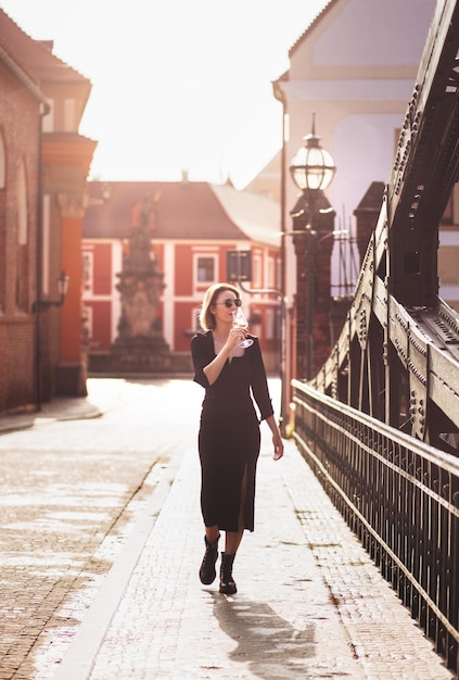 Attraktives, glückliches Mädchen in einem schwarzen Kleid geht mit einem Glas Wein auf einer Stadtstraße spazieren Glamouröse Lifestyle-Mode