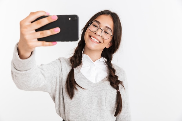 Attraktives glückliches Schulmädchen, das unifrom trägt, das isoliert über weißer Wand steht und ein Selfie macht