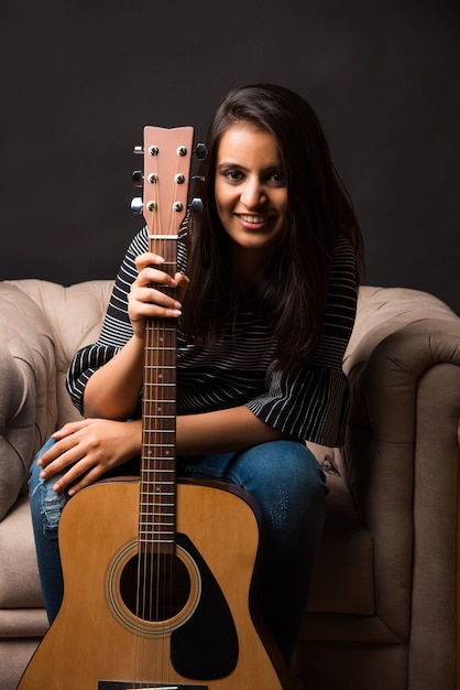 Attraktives indisches asiatisches junges Mädchen, das Gitarre spielt oder hält, während es zu Hause auf dem Sofa oder der Couch sitzt