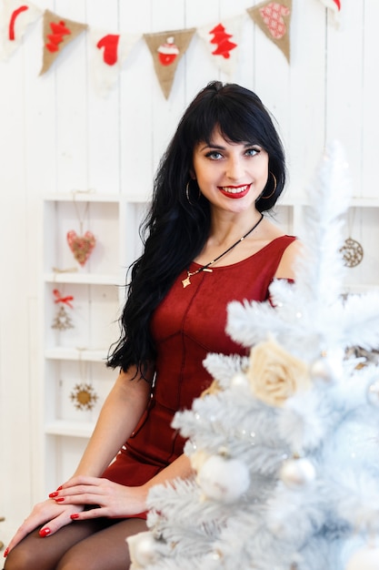 Attraktives junges Brunettemädchen im roten Kleid, das zuhause in einem Weihnachten sitzt, verzierte Innenraum.