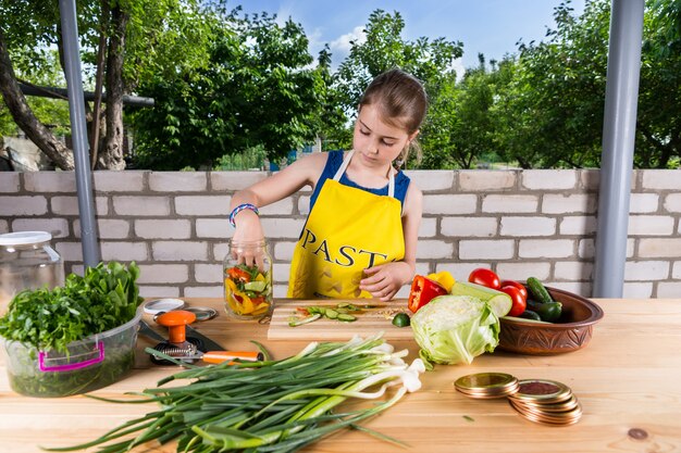 Attraktives junges Mädchen, das am Tisch im Garten im Garten steht und eine Auswahl an frischem Gemüse aus eigenem Anbau in Gläsern abfüllt