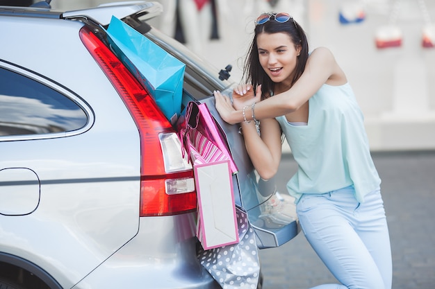 Attraktives junges Mädchen, das versucht, viele Einzelhandelsgeschäfte in ein Auto zu setzen. Frau, die Einkaufstaschen in ein Gepäck steckt. Junge Frau, die ihre Einkaufstaschen in einen Kofferraum schiebt.