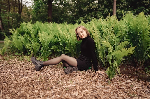 Attraktives junges Mädchen mit kurzen blonden Haaren sitzt im Sommer zwischen Farnen auf einer Wiese auf dem Boden