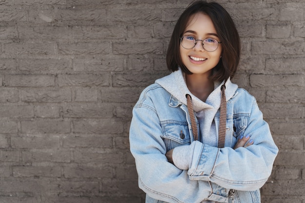 Attraktives junges Mädchen mit kurzen Haaren, Brille und Streetstyle-Kleidung, an eine Mauer gelehnt.