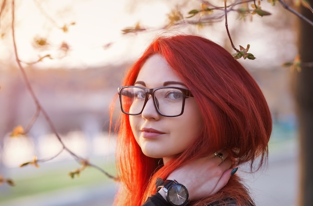 Attraktives junges Mädchen mit roten Haaren, das eine Brille trägt und bei Sonnenaufgang in der Nähe eines Baumes posiert