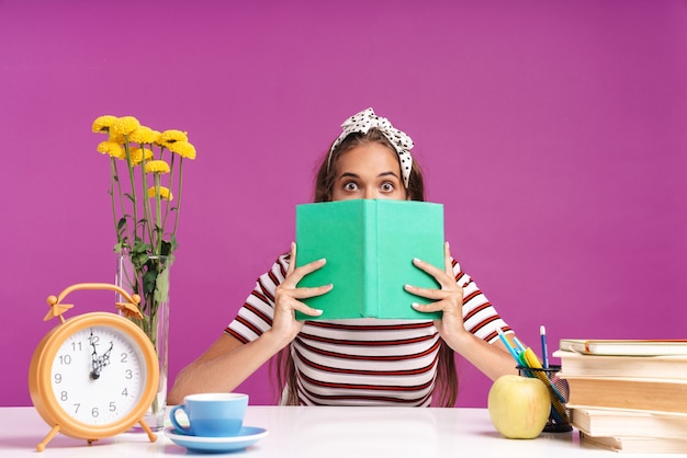 Attraktives junges Mädchen sitzt am Schreibtisch isoliert über rosa Wand, macht Hausaufgaben und bedeckt Gesicht mit Buch