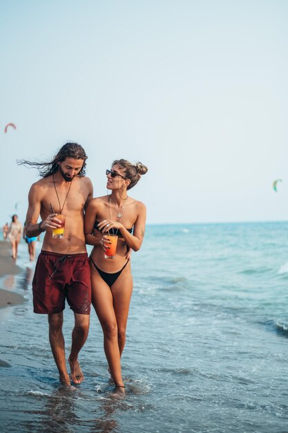 Attraktives junges Paar mit Alkoholcocktails, die am Strand spazieren gehen