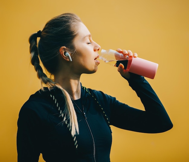 Attraktives kaukasisches Mädchen auf gelb isoliertem Hintergrund mit drahtlosen Kopfhörern im Sportanzug machen Training laufen und Trinkwasser