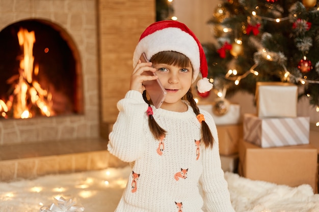 Attraktives Kind, das weißen Pullover und Weihnachtsmannhut spricht Telefon im festlichen Raum trägt