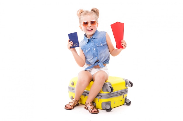 Attraktives kleines Mädchen im blauen Hemd, in den weißen kurzen Hosen und in der Sonnenbrille sitzen auf dem gelben Koffer