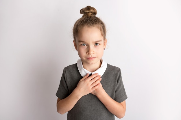 Attraktives kleines Mädchen in einem strengen Schulkleid, steht auf weißem Hintergrund, gestikuliert mit den Händen, bewundert einen glücklichen Ausdruck auf ihrem Gesicht. Foto in hoher Qualität