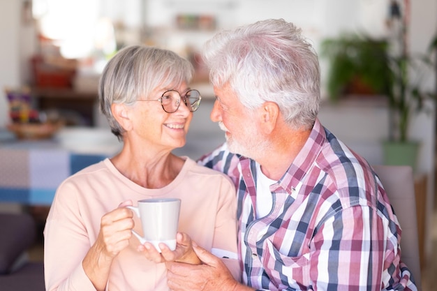 Attraktives lächelndes älteres Paar, das sich umarmt und in die Augen schaut, während die Frau eine Tasse Kaffee hält Starke Beziehung zwischen zwei kaukasischen Senioren, die sich wohlfühlen