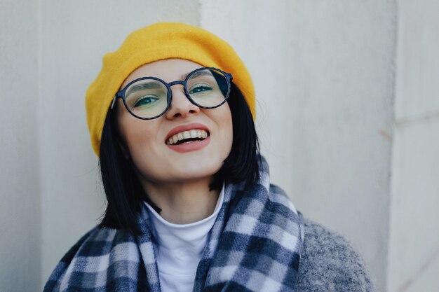 Attraktives lächelndes junges Mädchen in den Gläsern im Mantel und im gelben Barett auf einem einfachen hellen Hintergrund