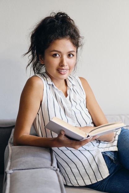 Attraktives, lächelndes, lässiges asiatisches Mädchen mit Buch, das glücklich in der Kamera auf ein modernes Zuhause blickt