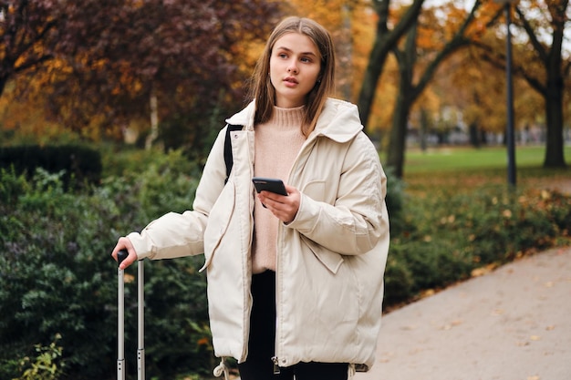 Attraktives lässiges Mädchen in Daunenjacke mit Handy, das nachdenklich im Stadtpark wegschaut