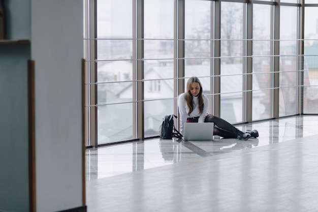 Attraktives Mädchen, das auf Boden mit Laptop und Dingen im Flughafenterminal oder im Büro sitzt. Reiseatmosphäre oder alternative Arbeitsatmosphäre.