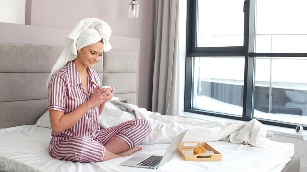 Attraktives Mädchen, das auf dem Bett sitzt, das an einem Laptop arbeitet. Selbstisolation, Quarantäne, Coronavirus