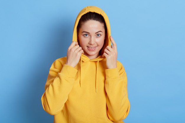 Attraktives Mädchen im gelben Sweatshirt mit Kapuze, die Kamera betrachtet und lächelt, steht gegen blaue Wand, hält Hände auf ihrer Kapuze, ist gut gelaunt und drückt positive Gefühle aus.