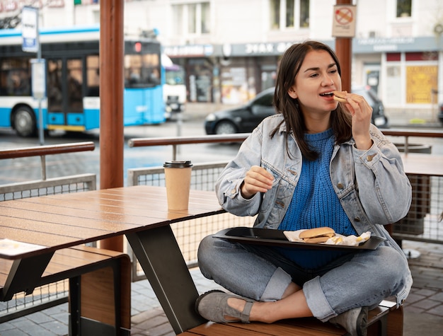 Attraktives Mädchen im lässigen Stil isst einen Burger mit Kaffee auf der Sommerterrasse eines Cafés