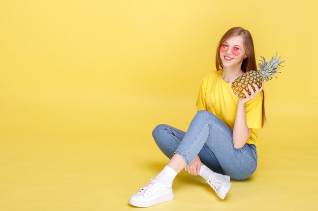 Attraktives Mädchen mit Ananas sitzt auf einer gelben Wand