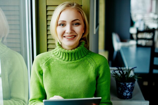 Attraktives Mädchen mit hellem Haar, das grünen Pullover trägt, sitzt im Café mit Tablette, freiberufliches Konzept, Online-Einkauf, Porträt.