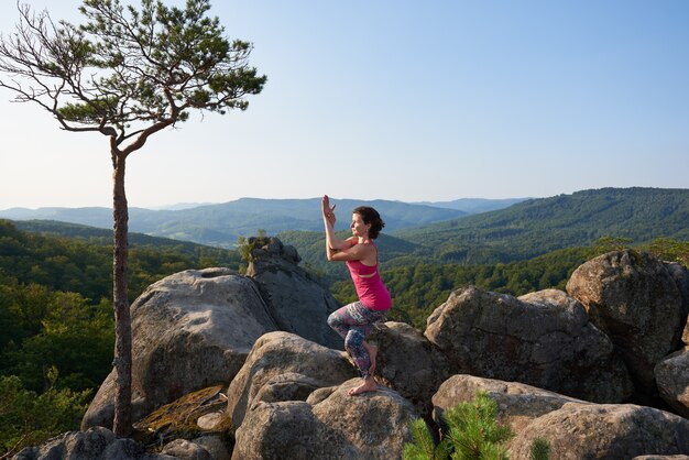 Attraktives Mädchen steht in komplizierter Yoga-Pose auf großem Felsen am warmen Sommertag