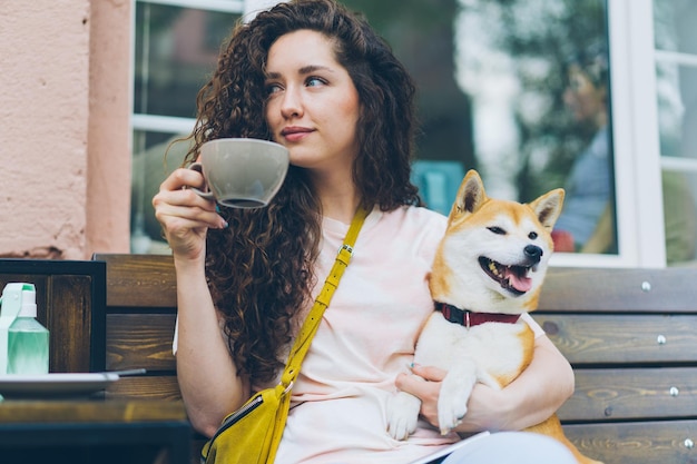 Attraktives Mädchen trinkt Tee im Freien im Café und streichelt den Hund, der im Café sitzt
