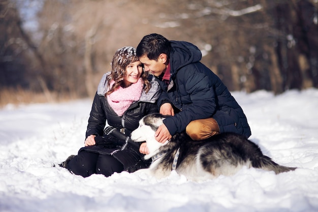 Attraktives Paar, das lächelt und Spaß im Winterpark mit ihrem siberian husky Hund hat.