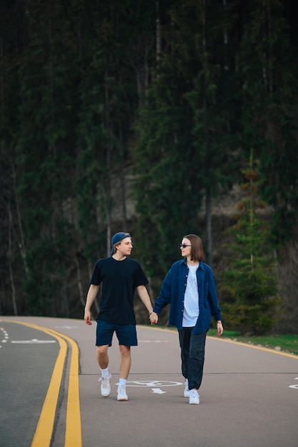 Attraktives Paar junger Leute, die auf einer asphaltierten Bergstraße im Wald spazieren und Händchen halten