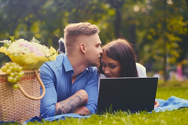Attraktives Paar liegt auf einer Decke auf einem Rasen und benutzt einen Laptop für ein Picknick.