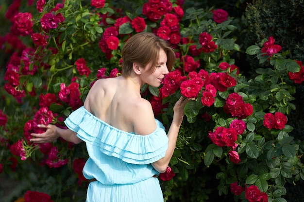 Attraktives Rothaarigemädchen mit der modischen Frisur, die das stilvolle Blaulichtkleid riecht blühende Rosen im Garten trägt