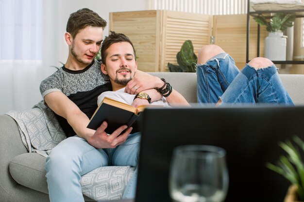 Attraktives schwules Paar, das Buch auf dem Sofa zu Hause im Wohnzimmer liest