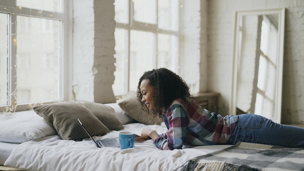 Attraktives Teenager-Mädchen, das mit Laptop lacht, um soziale Medien zu teilen, die zu Hause im Bett liegen