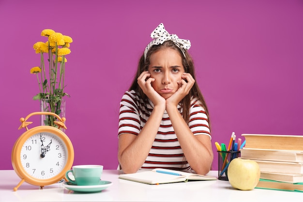 Attraktives verärgertes junges Mädchen, das isoliert über rosa Wand am Schreibtisch sitzt und Hausaufgaben macht